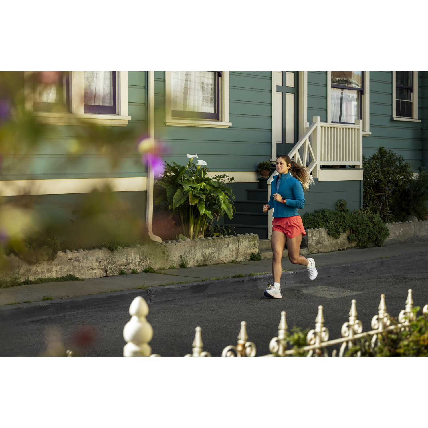 A woman in a blue top and red shorts runs on a street in front of a house.