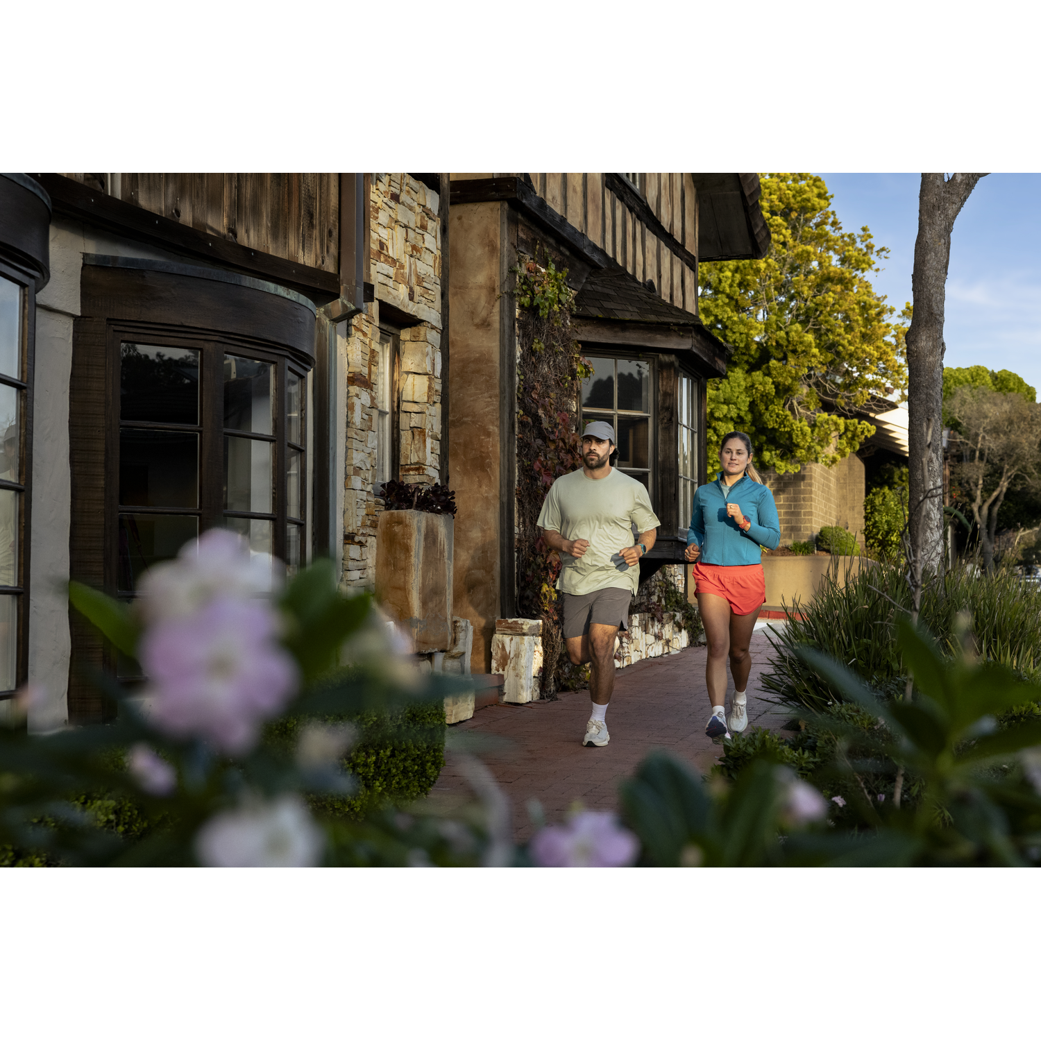 A man and woman jog on a sidewalk in front of a stone house.