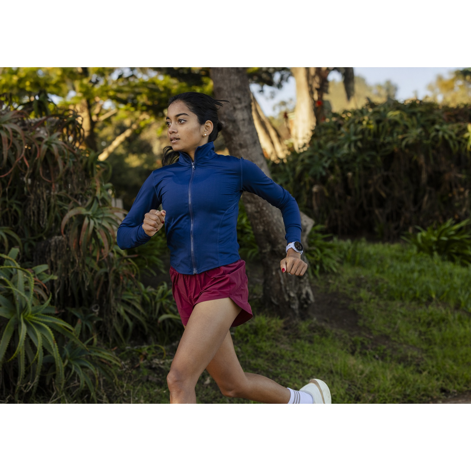 Une femme en veste bleue et short rouge court dehors dans la verdure.
