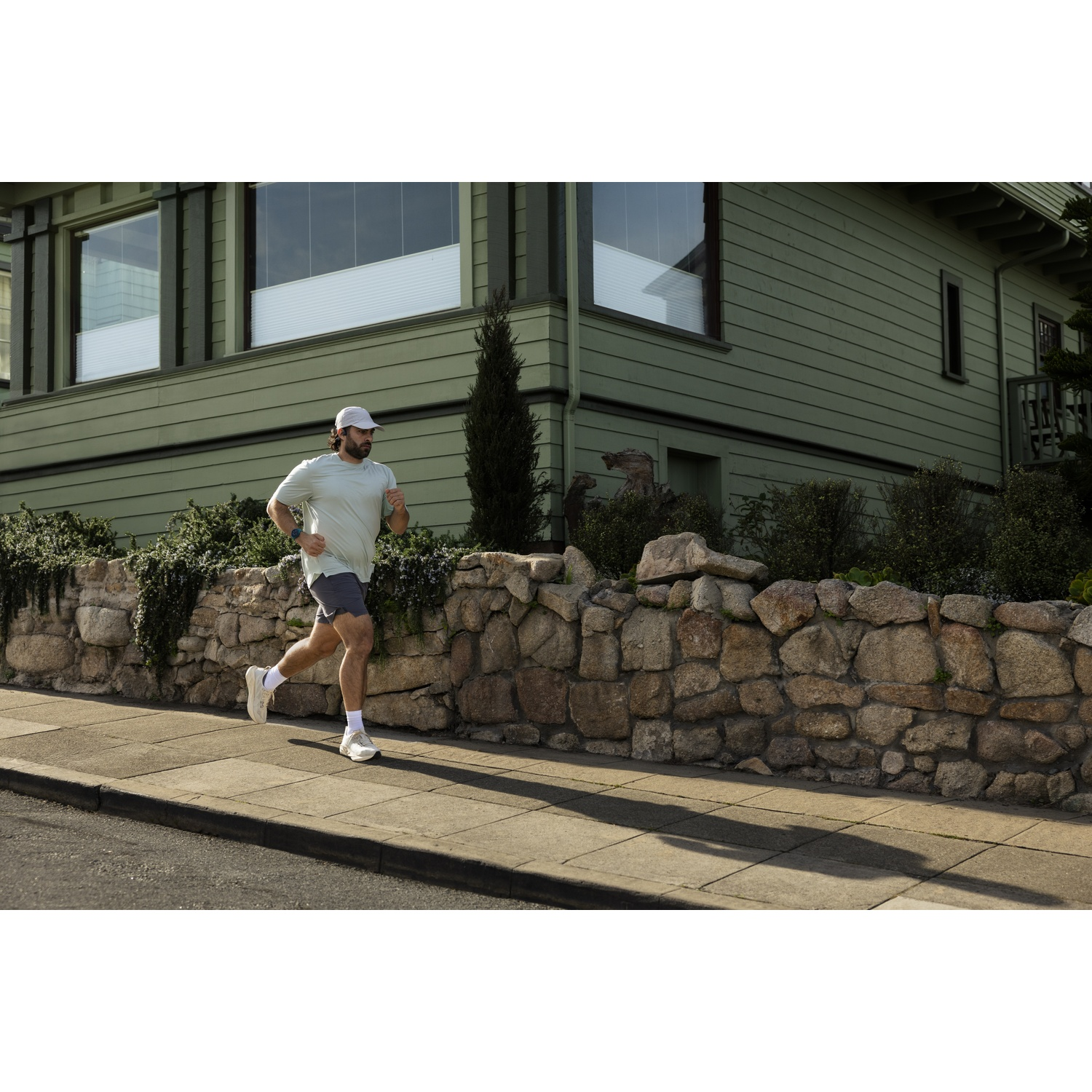 Man in light blue shirt and grey shorts runs on sidewalk by stone wall.