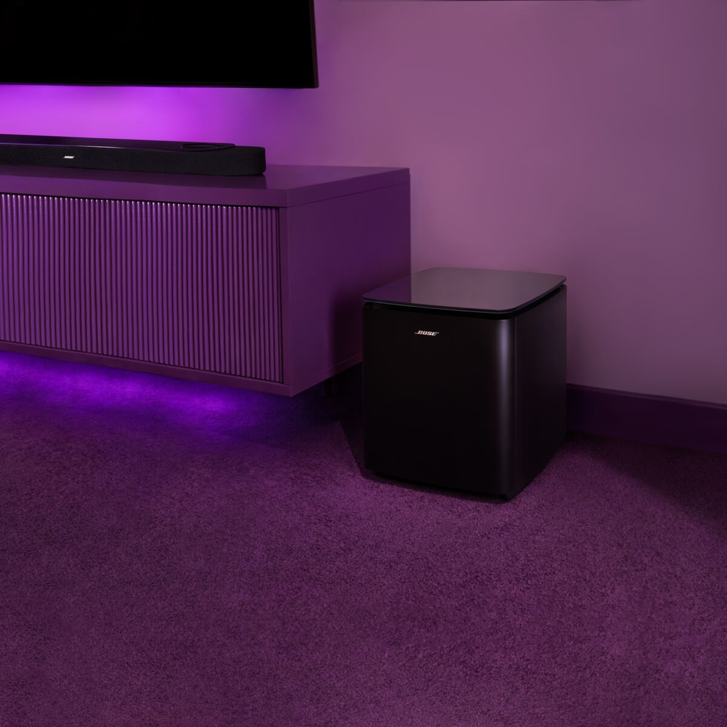 A black Bose subwoofer sits on a purple carpet next to a cabinet with a soundbar.