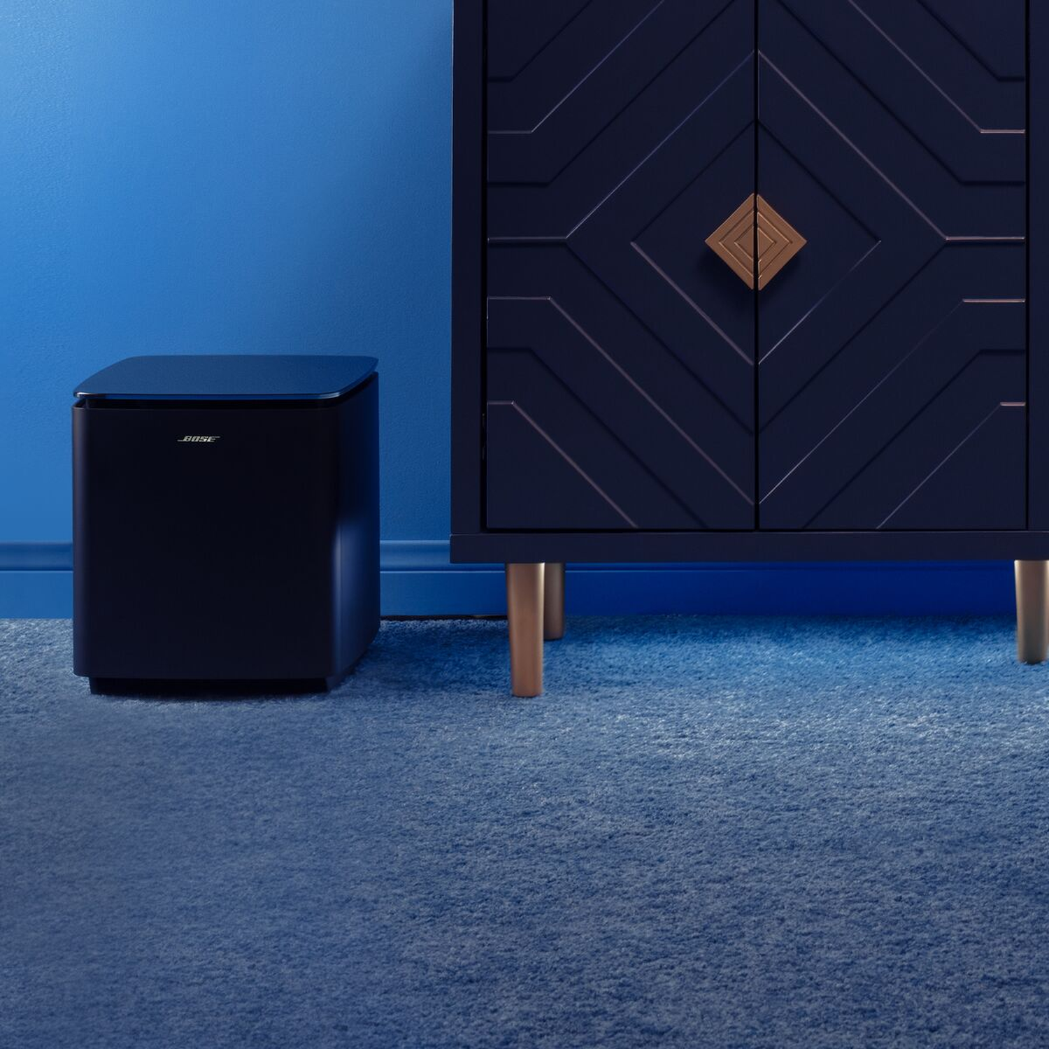 A dark Bose subwoofer sits next to a navy blue cabinet on a blue carpet.