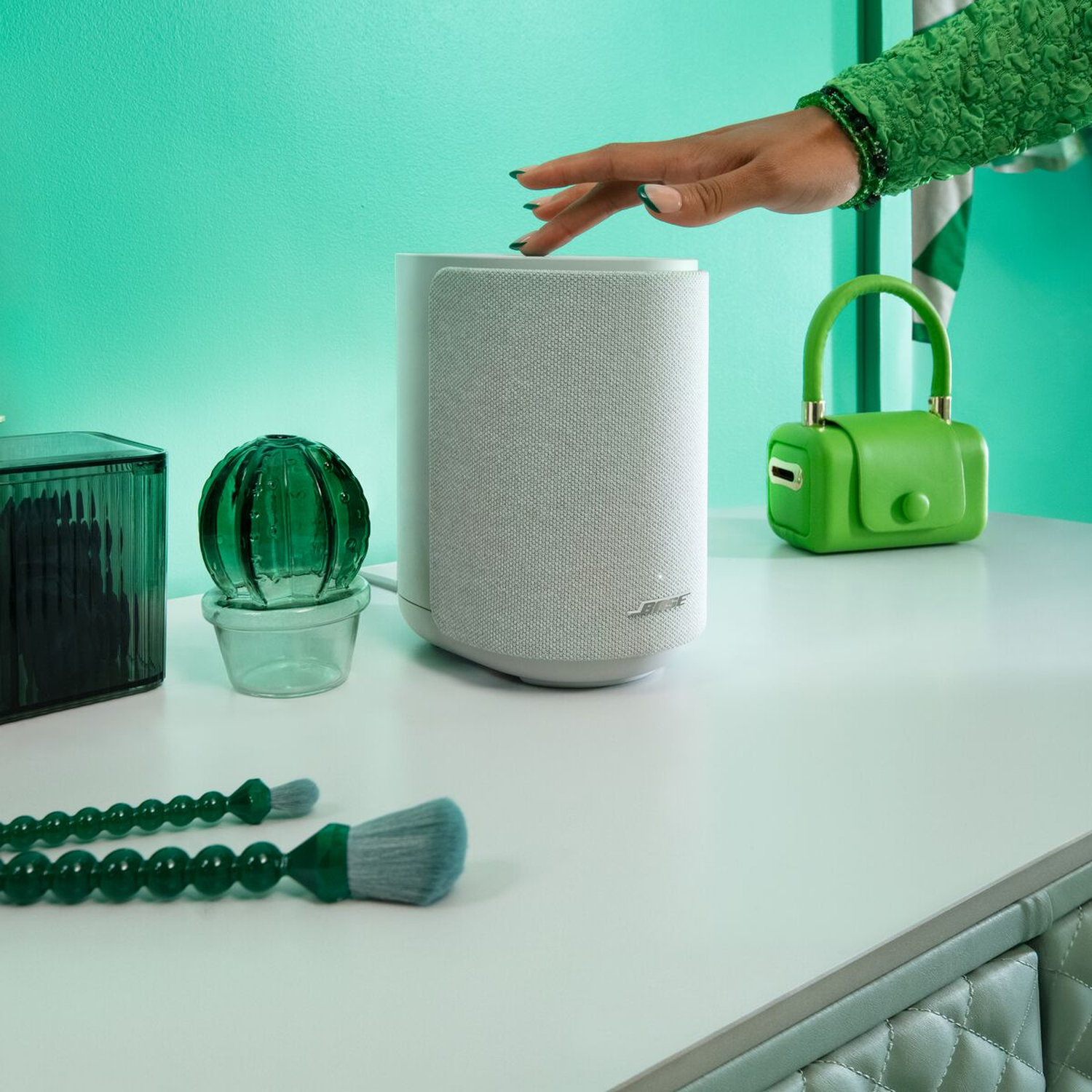 Een hand raakt een witte Bose-speaker aan. Groene accessoires omvatten een tas, vaas en kwasten.