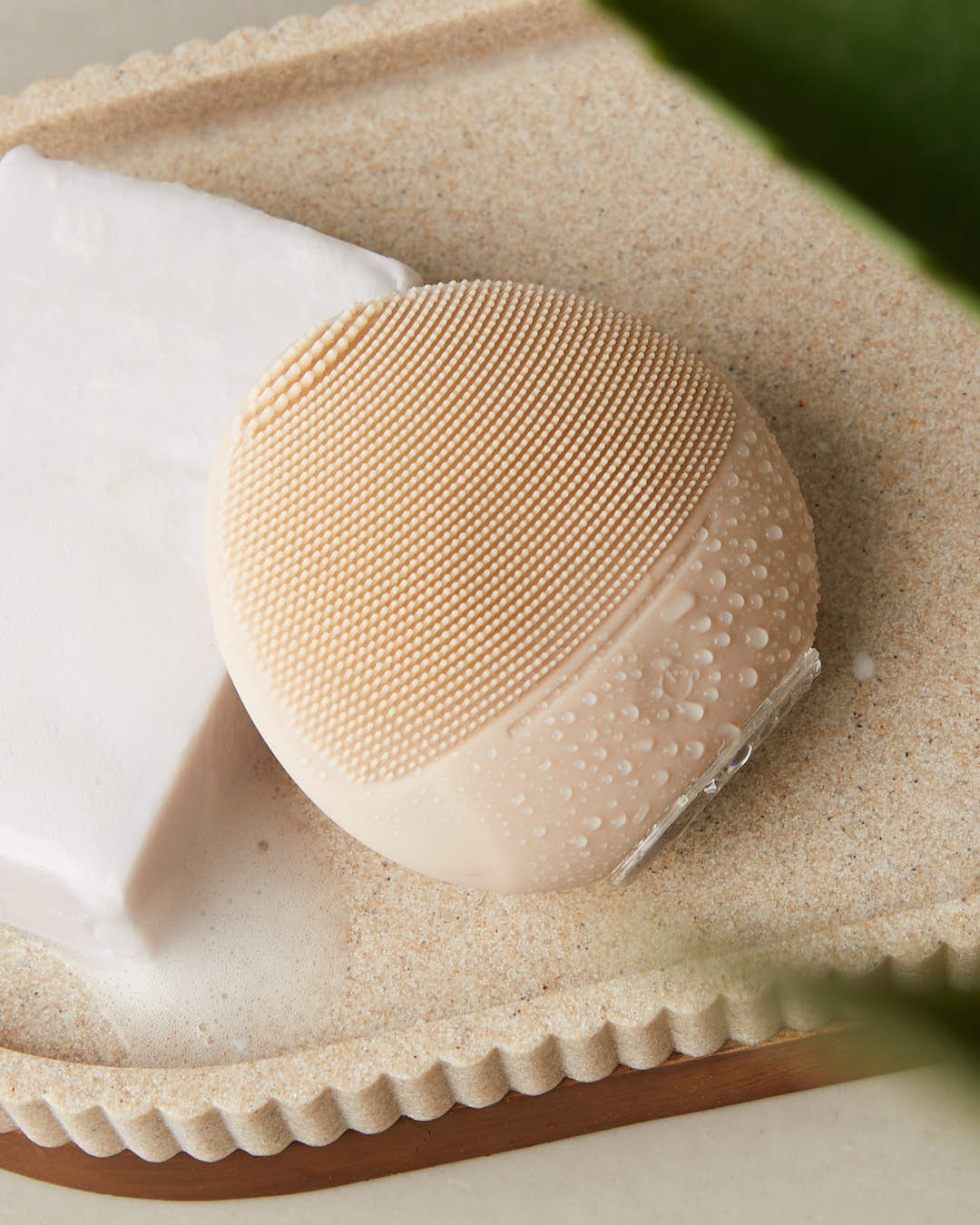 Un cepillo limpiador facial de silicona beige con gotas de agua se asienta en una bandeja texturizada junto a jabón blanco.