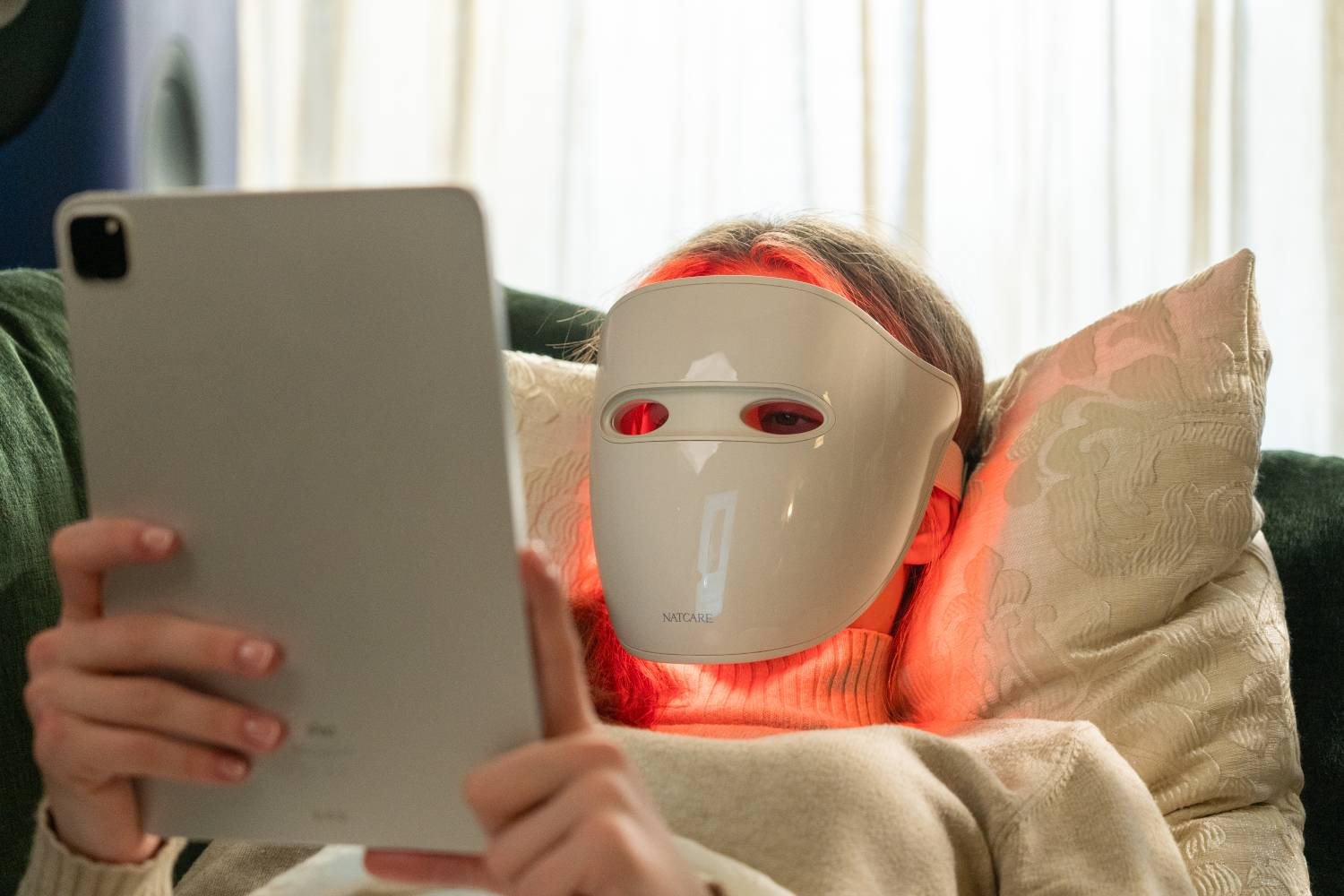 Persona con mascarilla LED y tablet en el sofá.