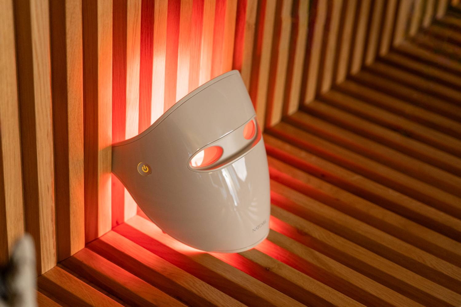 Una mascarilla facial LED beige con luces naranjas brillantes está sobre una superficie de madera acanalada.