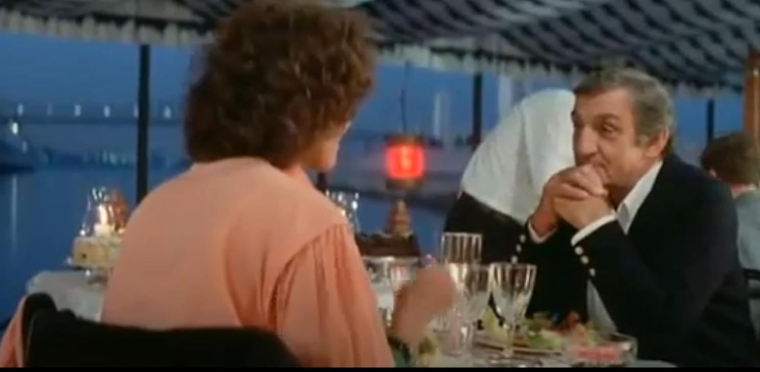 Ein Mann und eine Frau sitzen an einem Tisch mit Blick auf das Wasser. Der Mann trägt einen schwarzen Anzug.