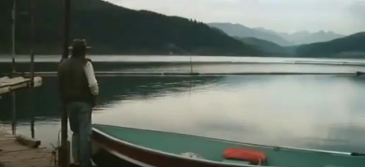 Ein Mann in Hut und Weste steht am Steg neben einem Boot auf einem ruhigen See.