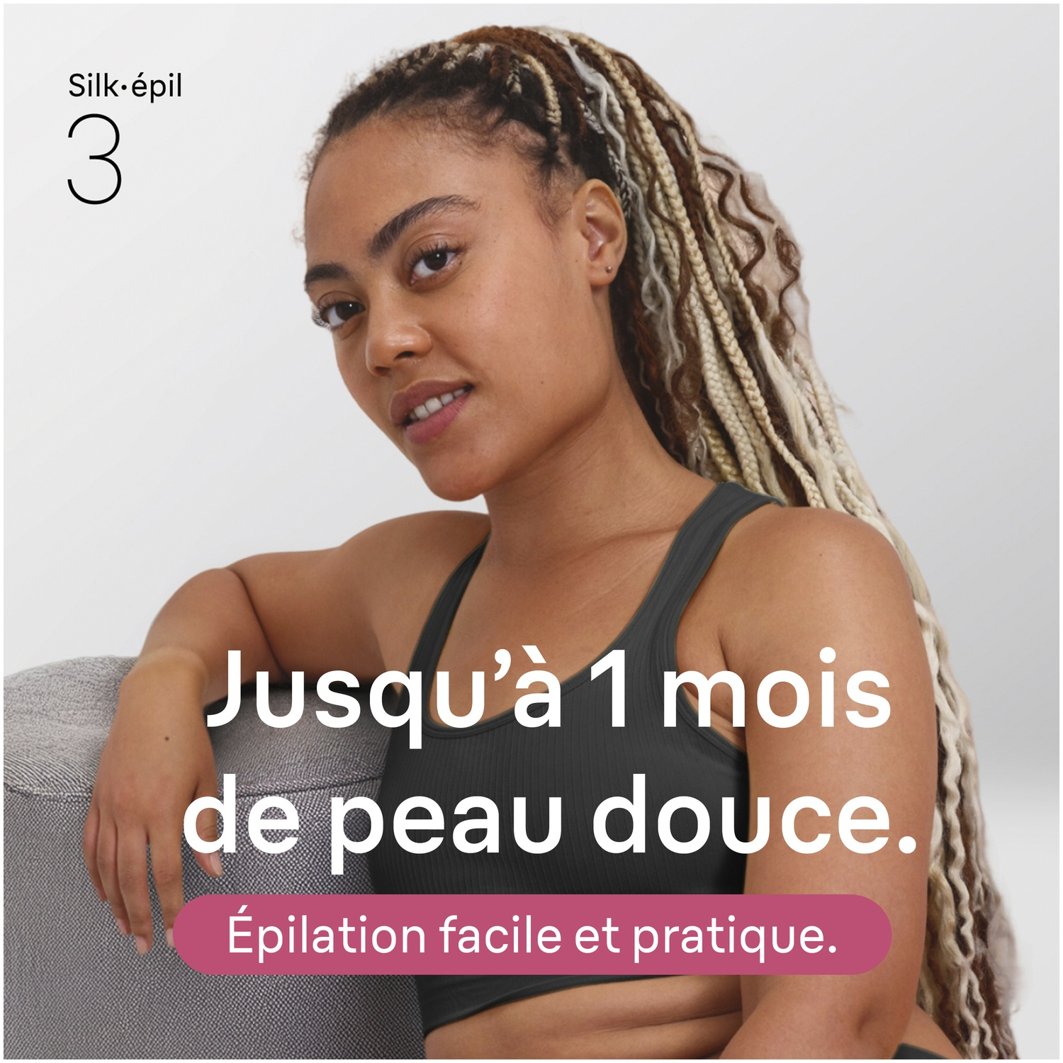 Une femme aux cheveux tressés assise sur un canapé gris. Texte sur peau douce.
