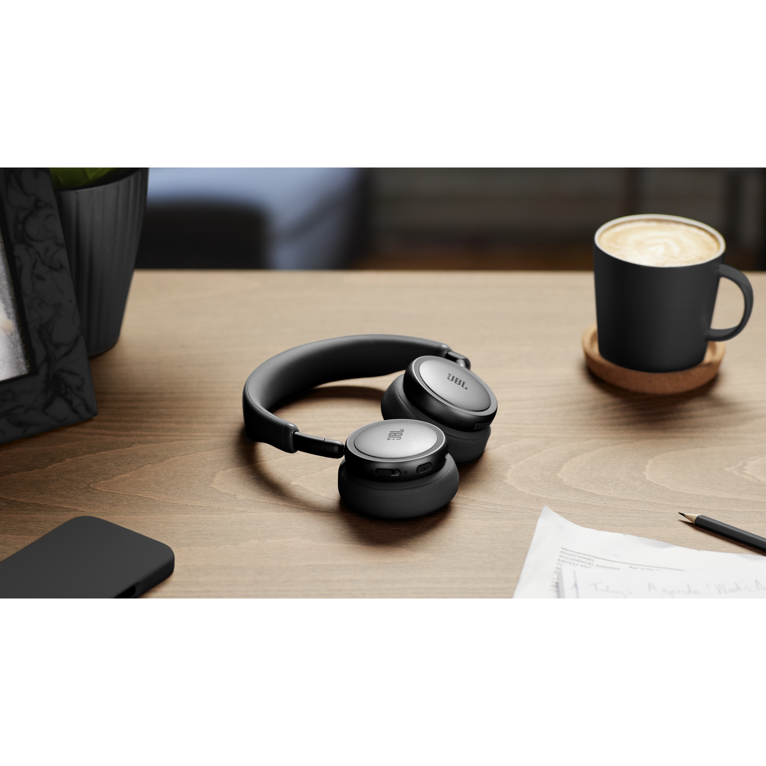Black JBL headphones, black mug of coffee, and a smartphone on a wooden desk.