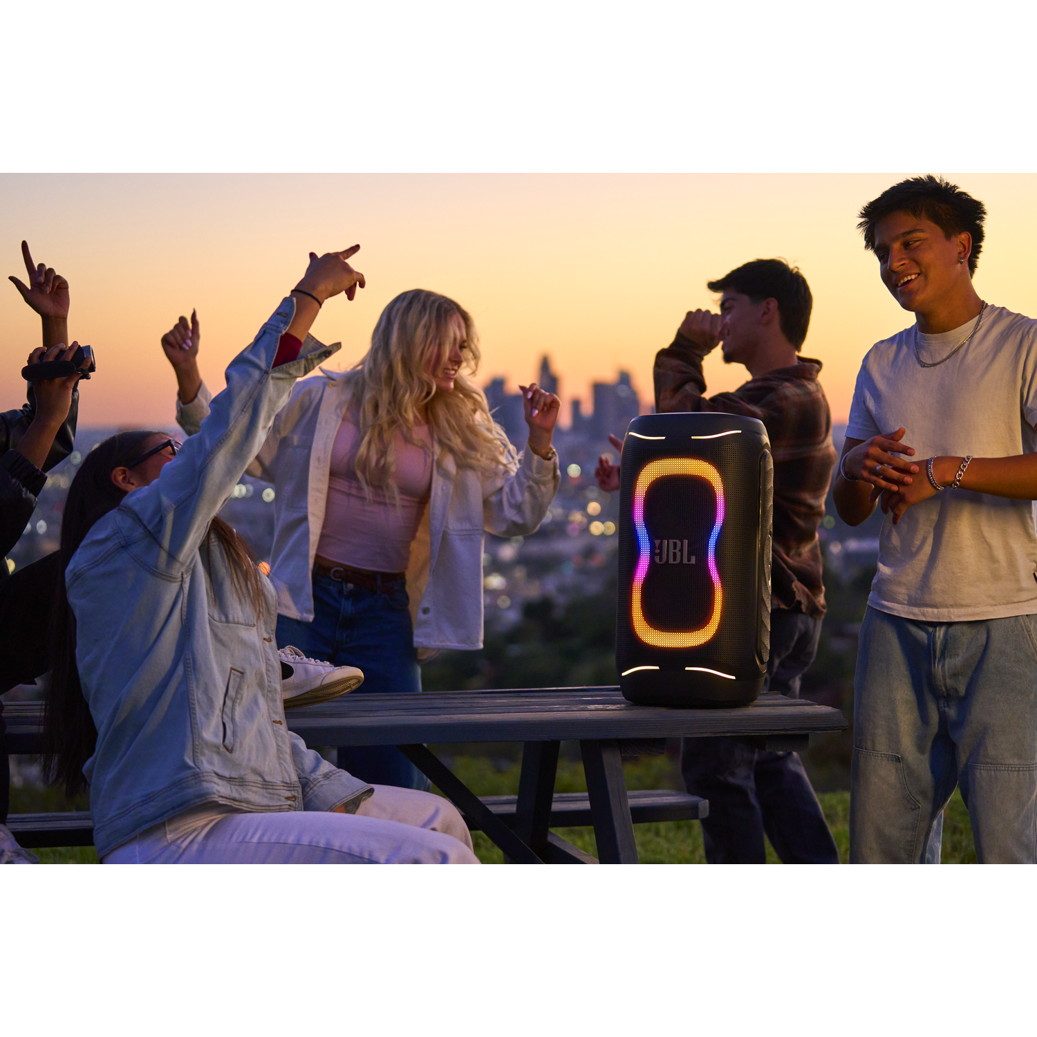Teens dance around a glowing JBL speaker at sunset.