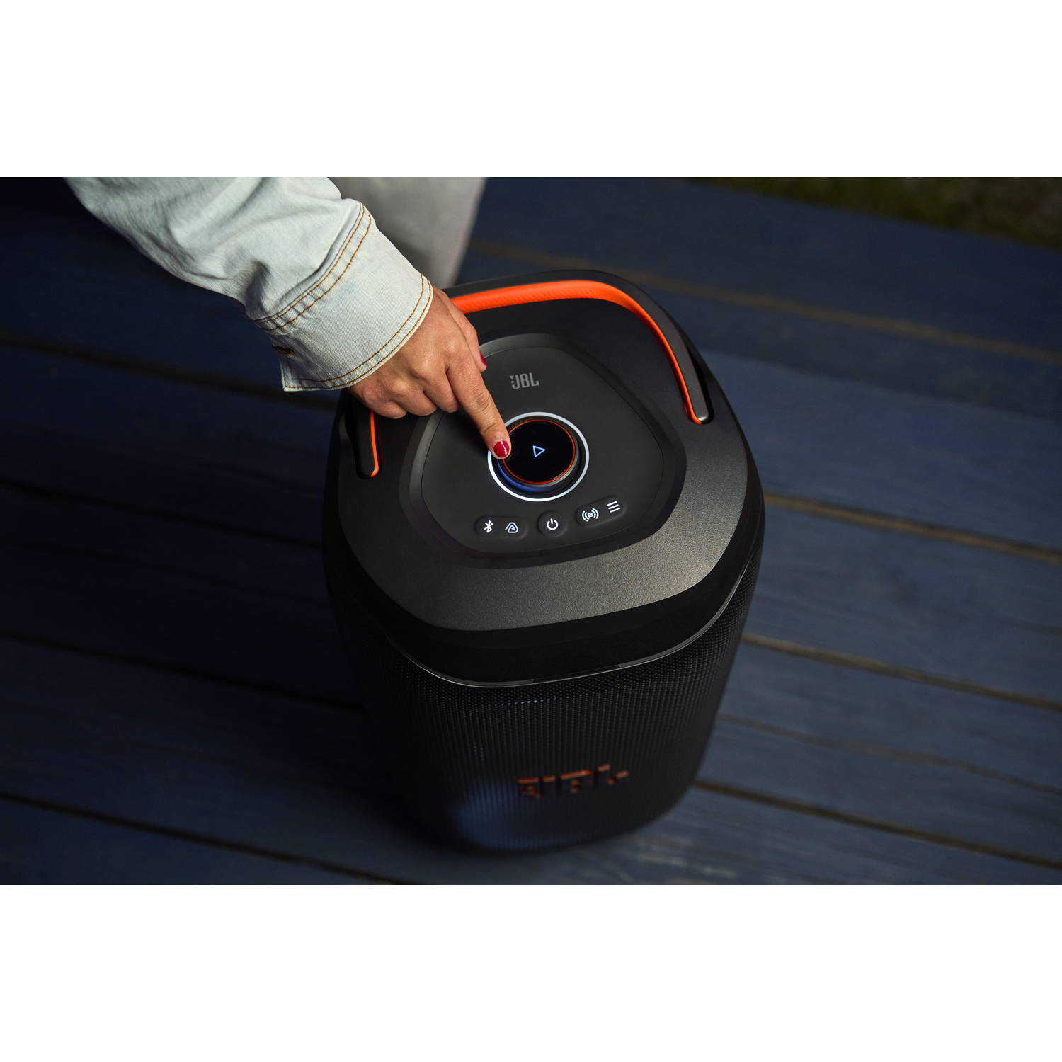 A finger presses play on a black JBL speaker with an orange handle on a blue wooden deck.