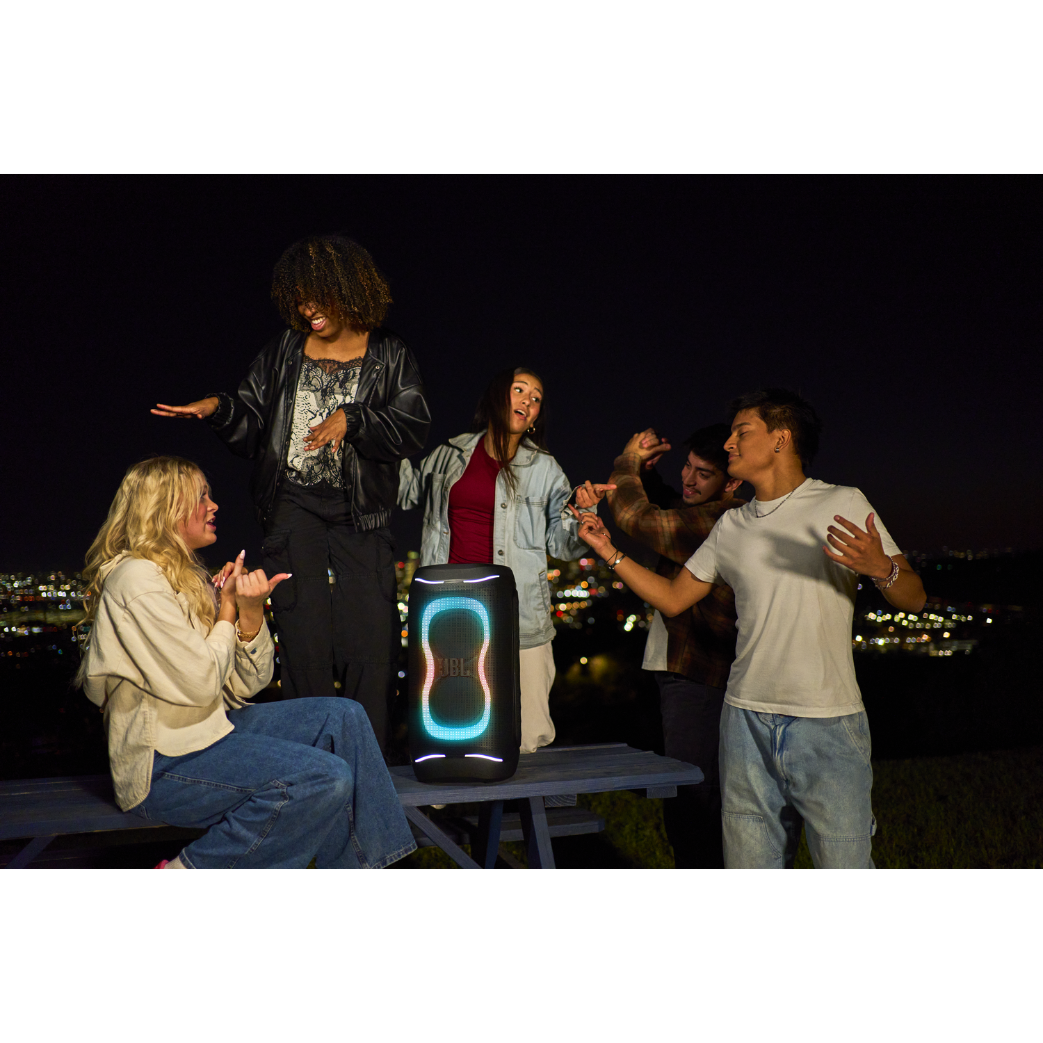 Five friends dance around a JBL speaker at night with city lights in the background.