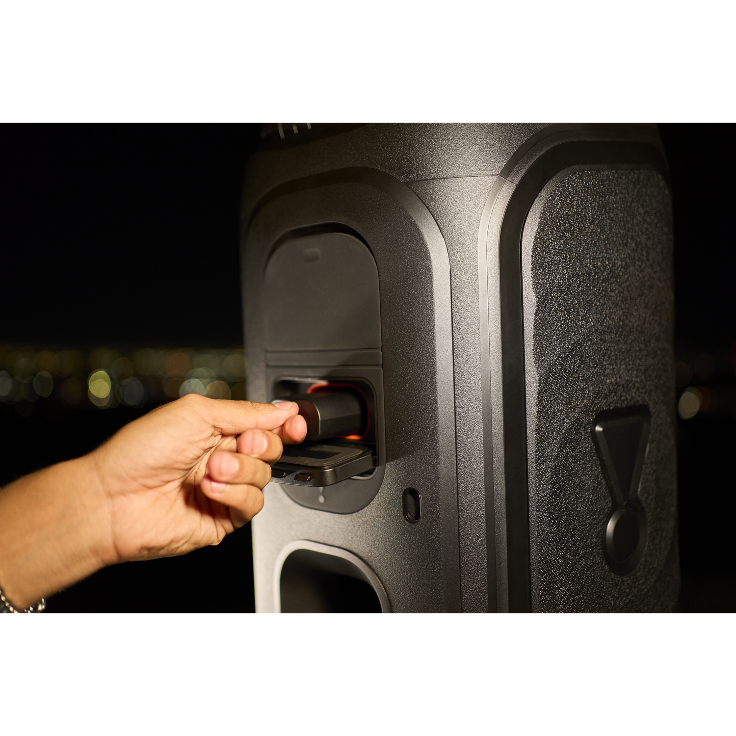Hand inserts USB stick into black speaker with lights in background.