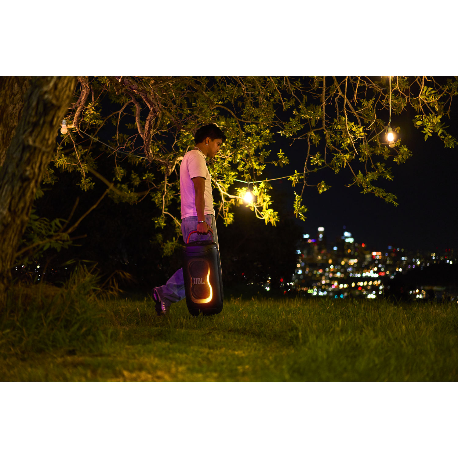 A man carries a glowing JBL speaker on a grassy hill at night.