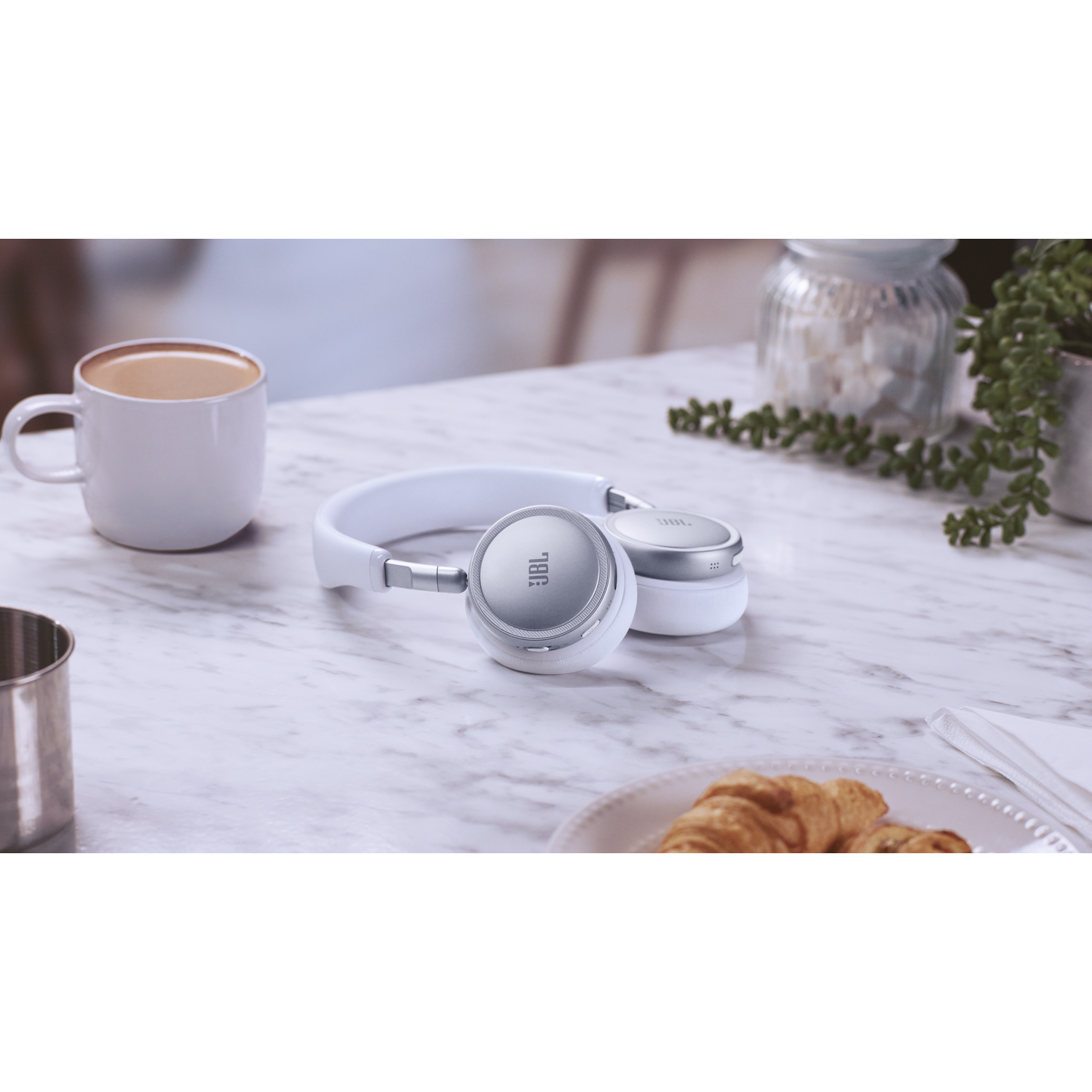 White headphones with silver accents on a marble table with coffee and croissant.