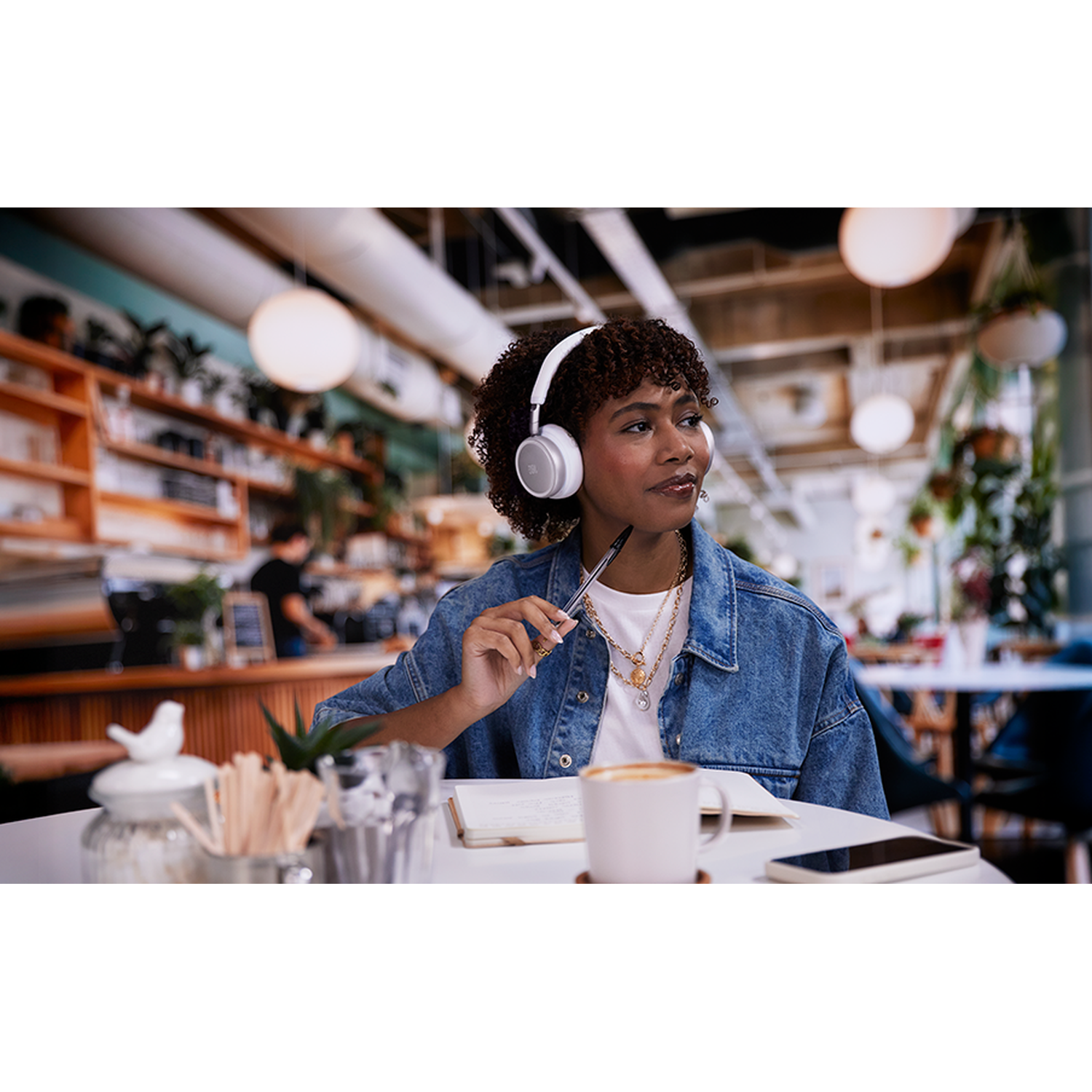 A woman wearing headphones sits in a cafe, holding a pen.