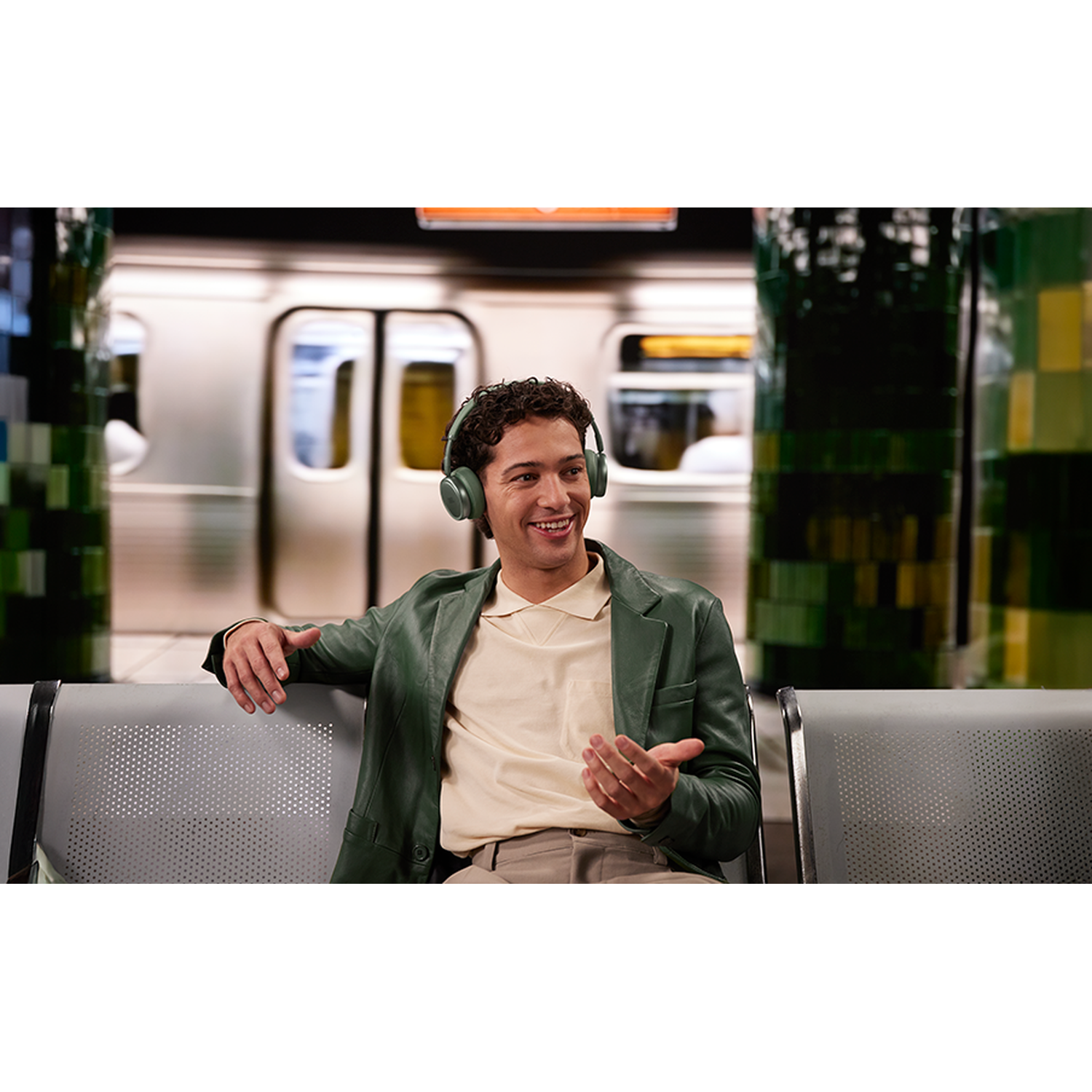 A man wearing headphones sits smiling in a subway station.