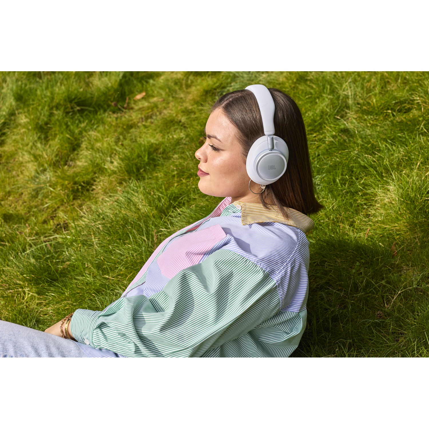 Une femme assise dans l'herbe portant des écouteurs blancs et une chemise rayée.