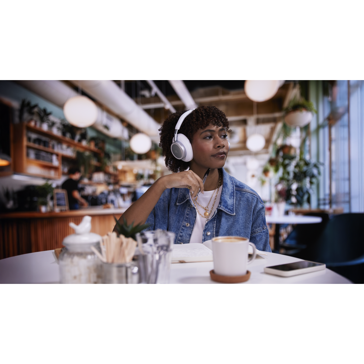 A woman wearing headphones sits in a cafe, looking thoughtfully out the window.
