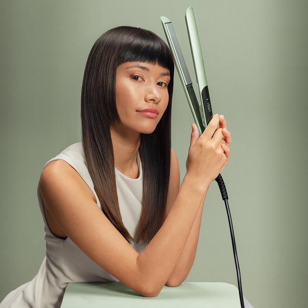 Una mujer con pelo largo sostiene una plancha verde sobre fondo verde pastel.