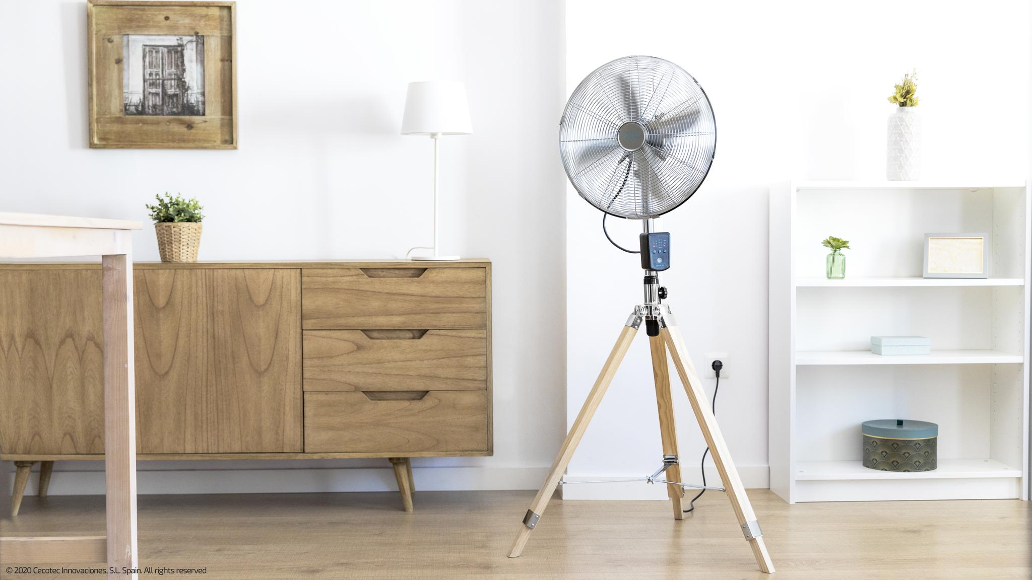 Un ventilador plateado sobre un trípode de madera está junto a un mueble de madera con una lámpara.
