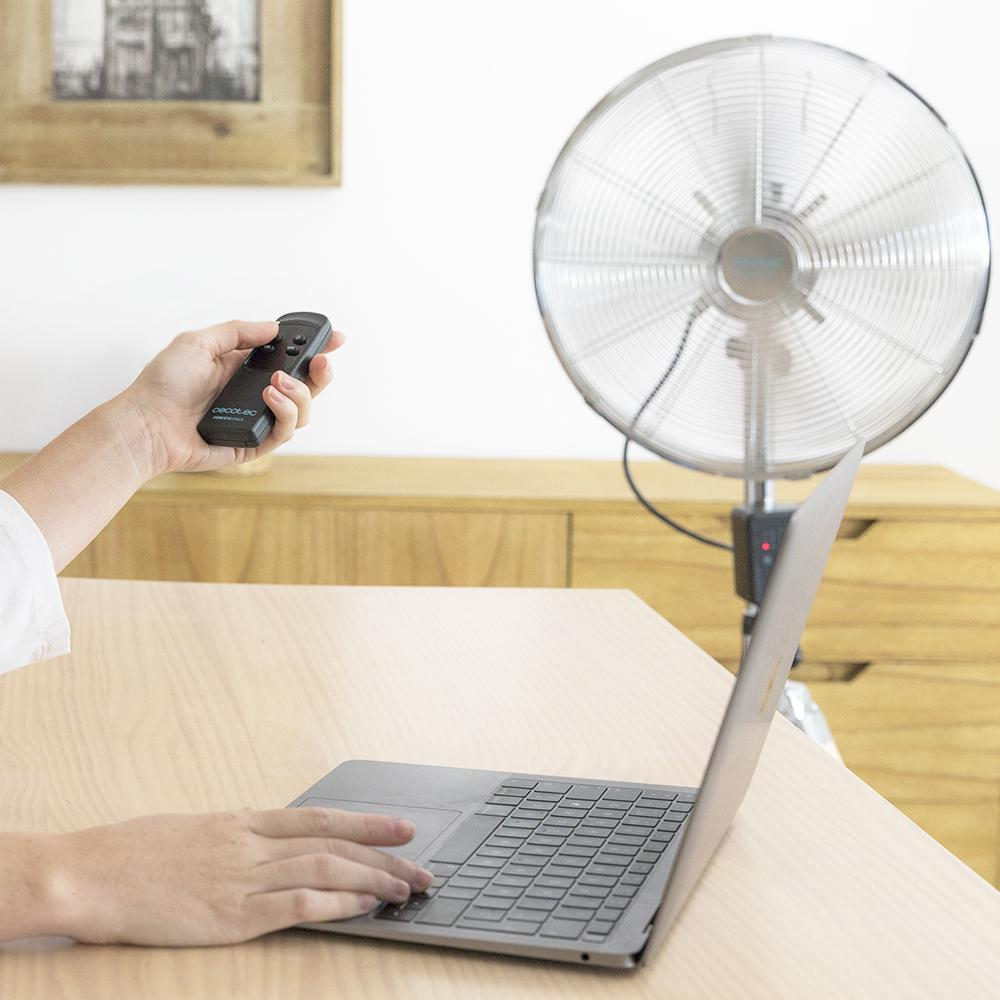 Una mano sostiene un control remoto, un portátil sobre un escritorio de madera, un ventilador al fondo.