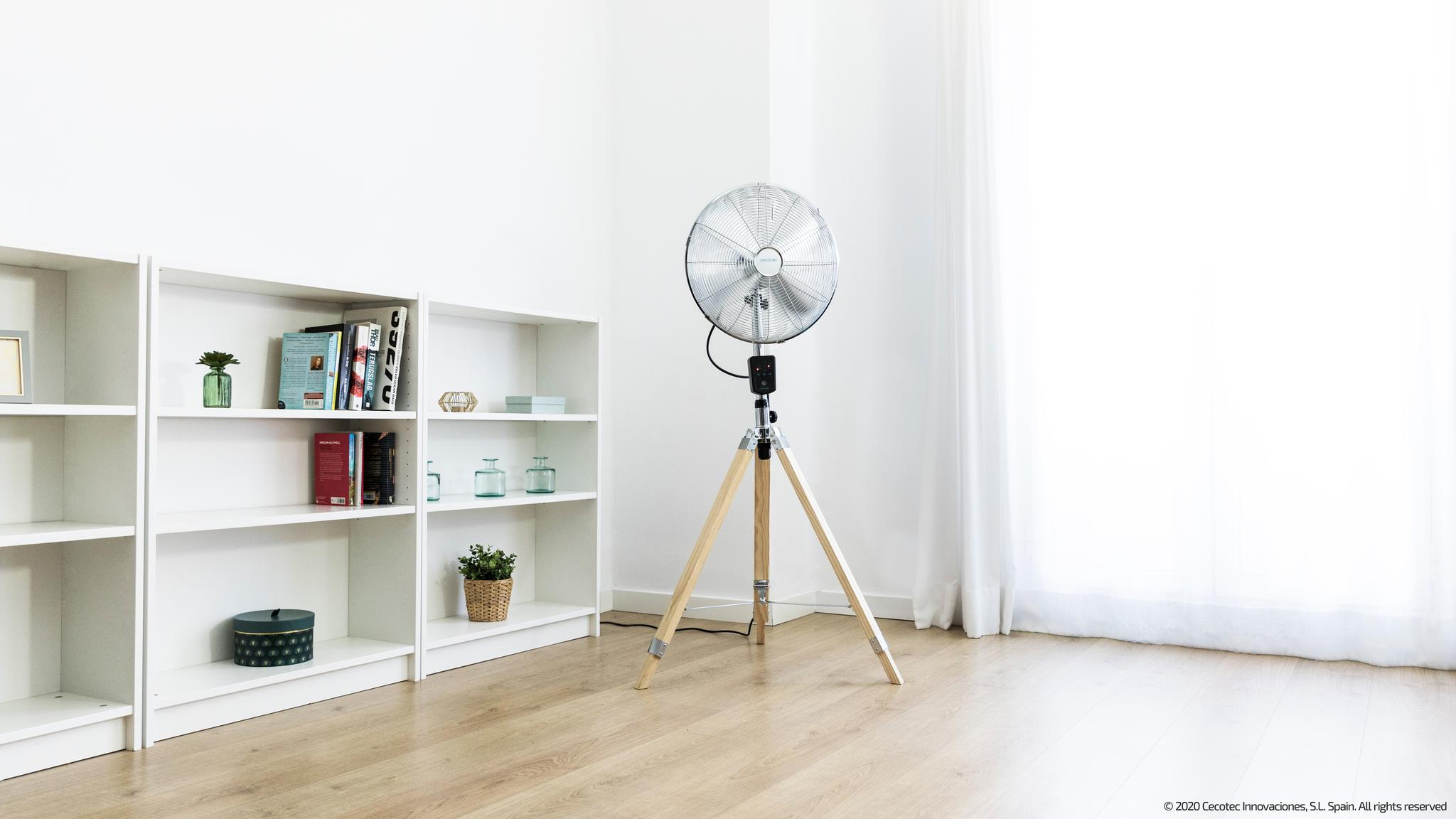 Un ventilador metálico sobre un trípode de madera junto a estanterías blancas con libros y decoración.