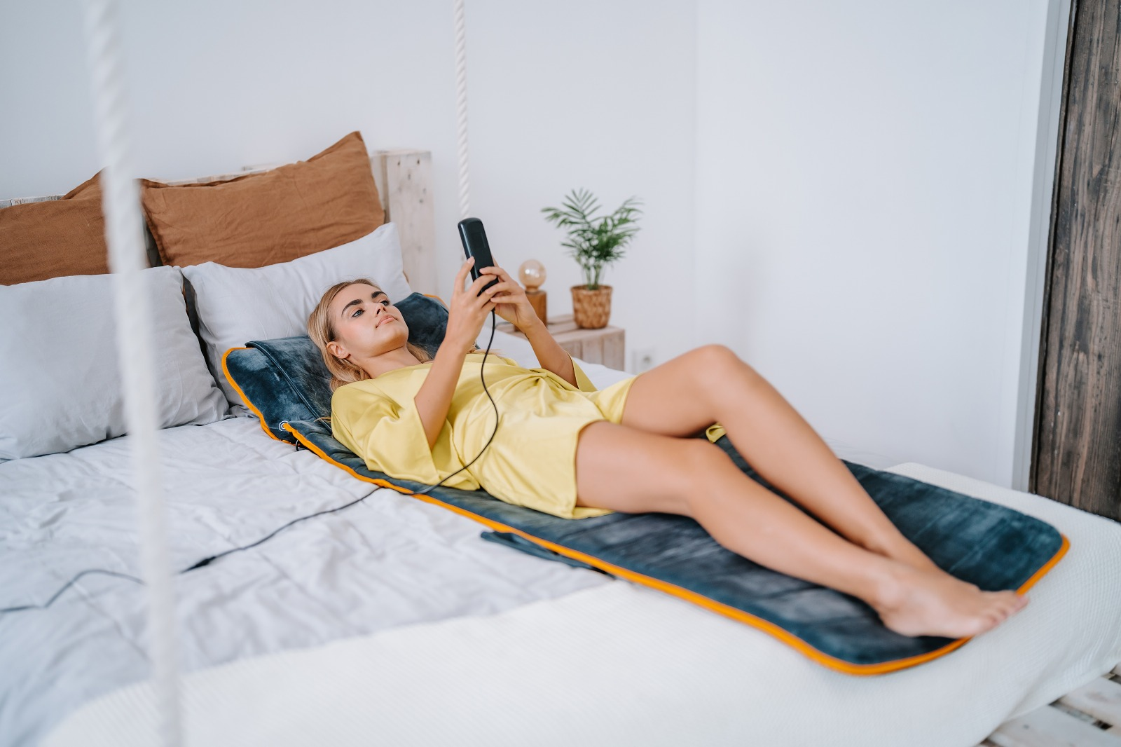 Kobieta w żółtej sukience leży na łóżku i patrzy na telefon.