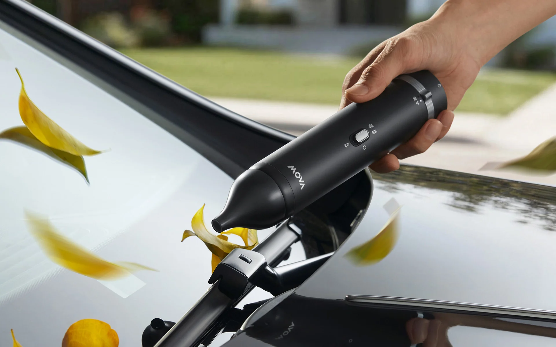 Hand hält schwarzen Mova-Sauger auf Auto-Windschutzscheibe mit gelben Blättern.