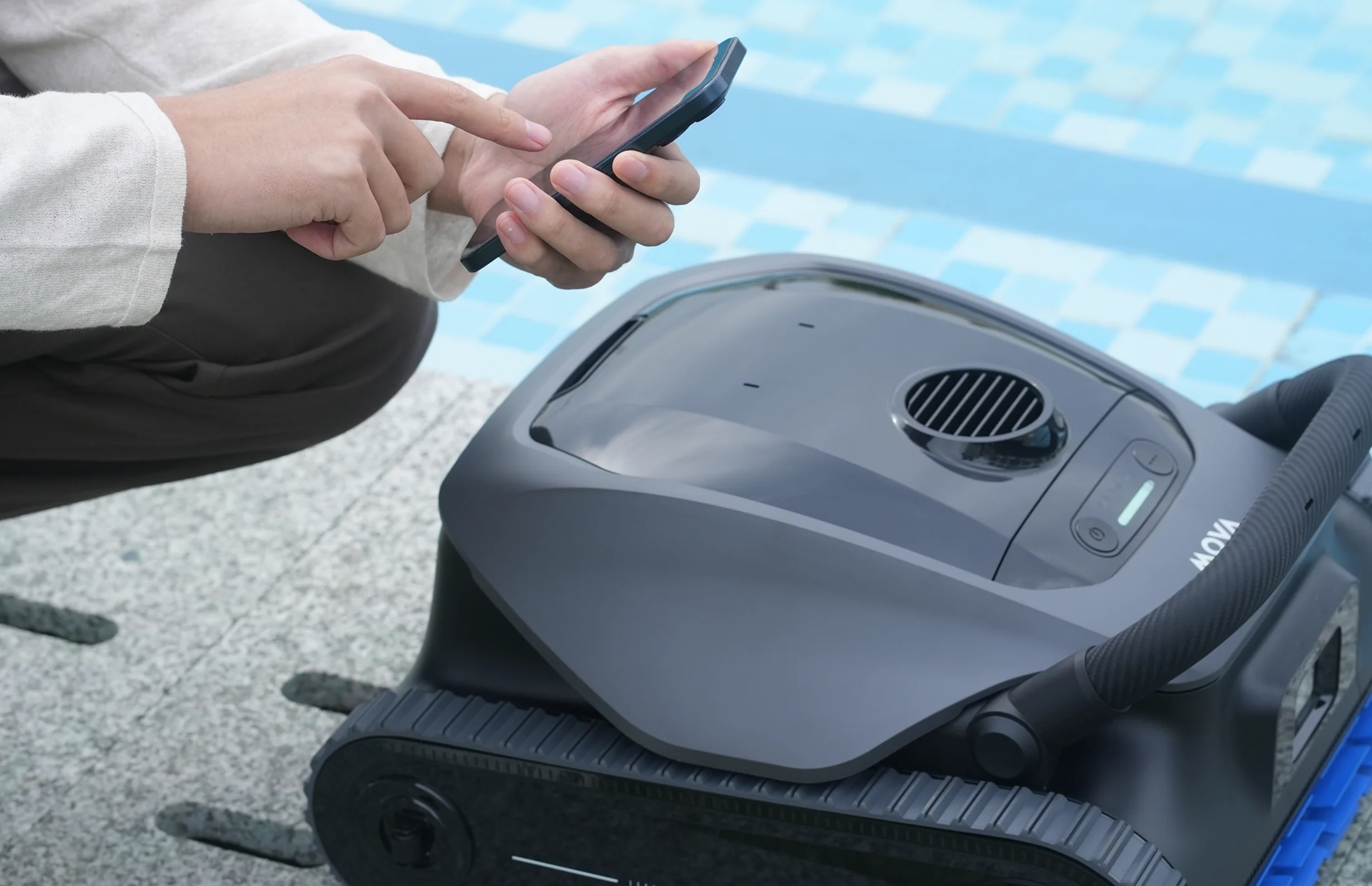 Person bedient Smartphone neben Poolroboter am Beckenrand.