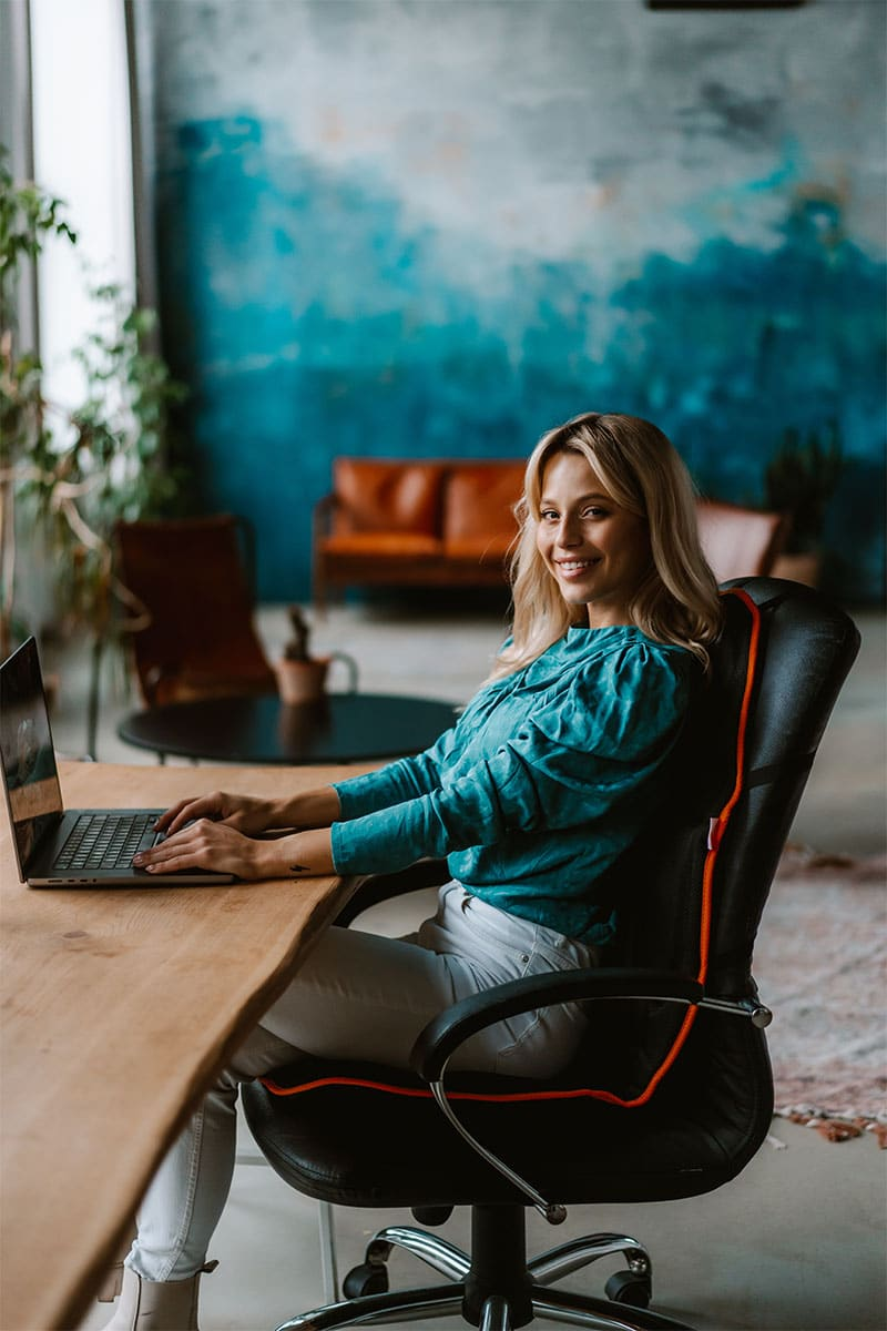 Blondynka siedzi przy drewnianym biurku i pisze na laptopie.