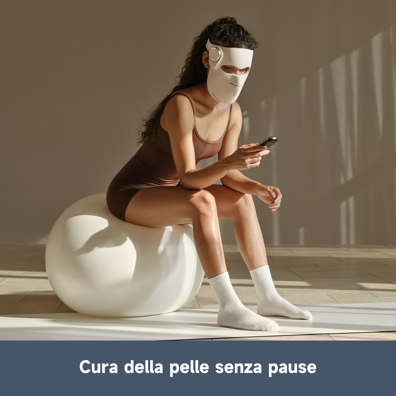 Donna con maschera facciale seduta su palla grande guarda il telefono.