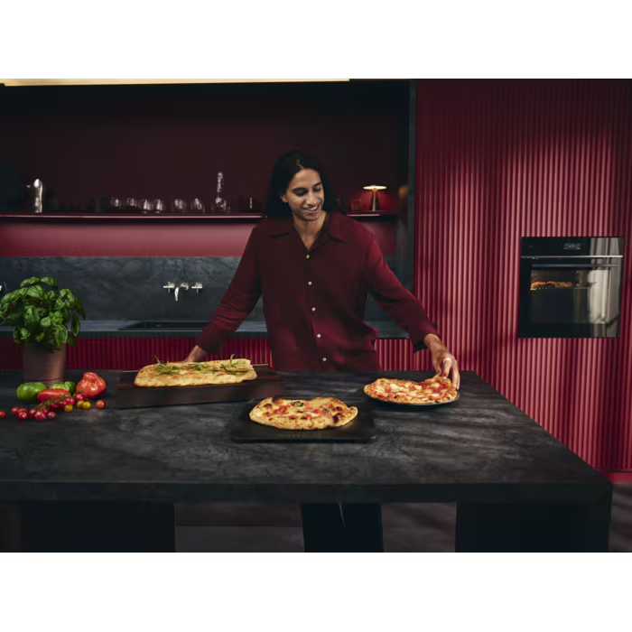 Egy piros inges nő áll egy asztalnál három pizzával. A háttérben sütő látható.