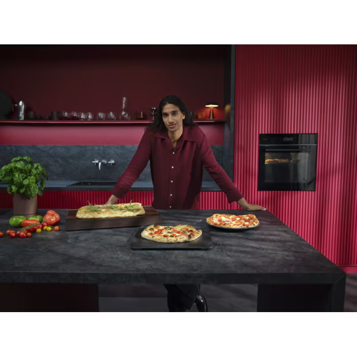 Egy férfi piros falú konyhában áll, három pizzát mutat be az asztalon.