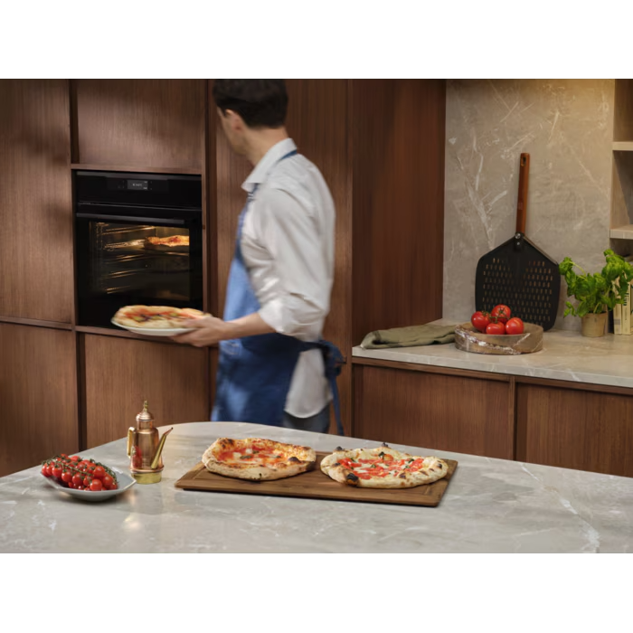 Egy kék kötényes férfi húz ki pizzát a sütőből. Két pizza van egy deszkán.