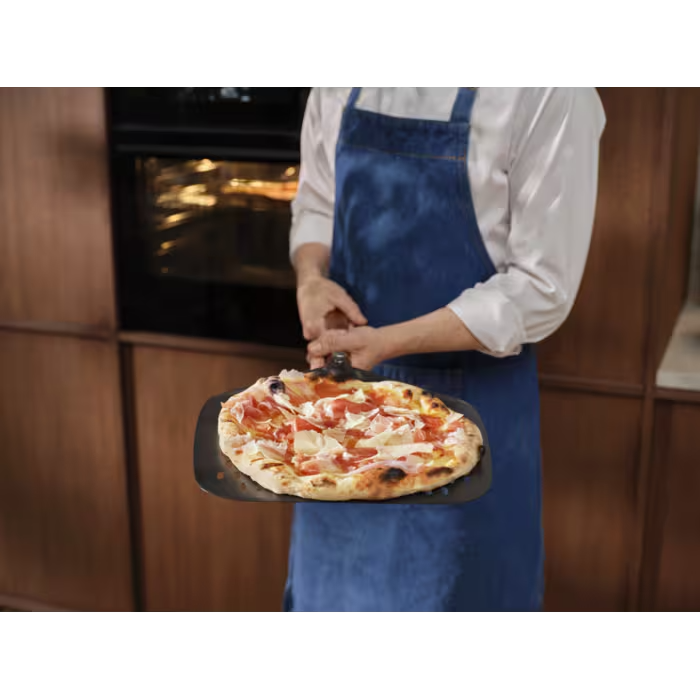 Egy kék kötényes szakács sonkás, paradicsomos pizzát tart pizzalapáton.