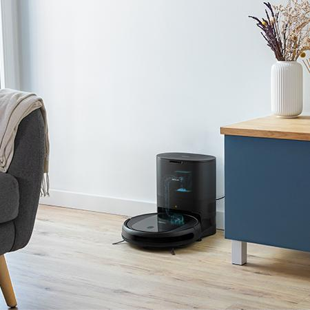 Un robot aspirador negro con estación de carga se sienta en un suelo de madera junto a un sillón gris.