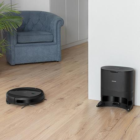 Un robot aspirador negro sobre suelo de madera cerca de sillón azul y base de carga.