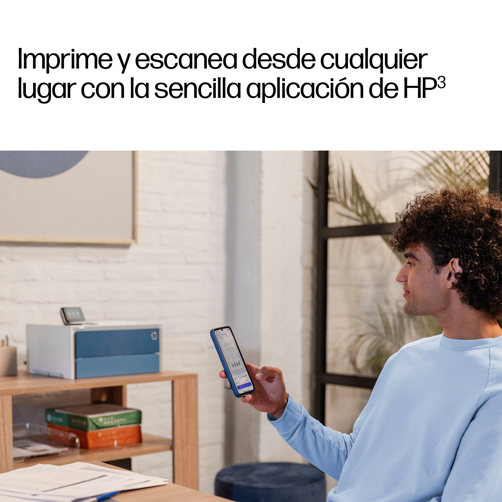 Hombre sostiene smartphone frente a impresora en escritorio, con plantas atrás.