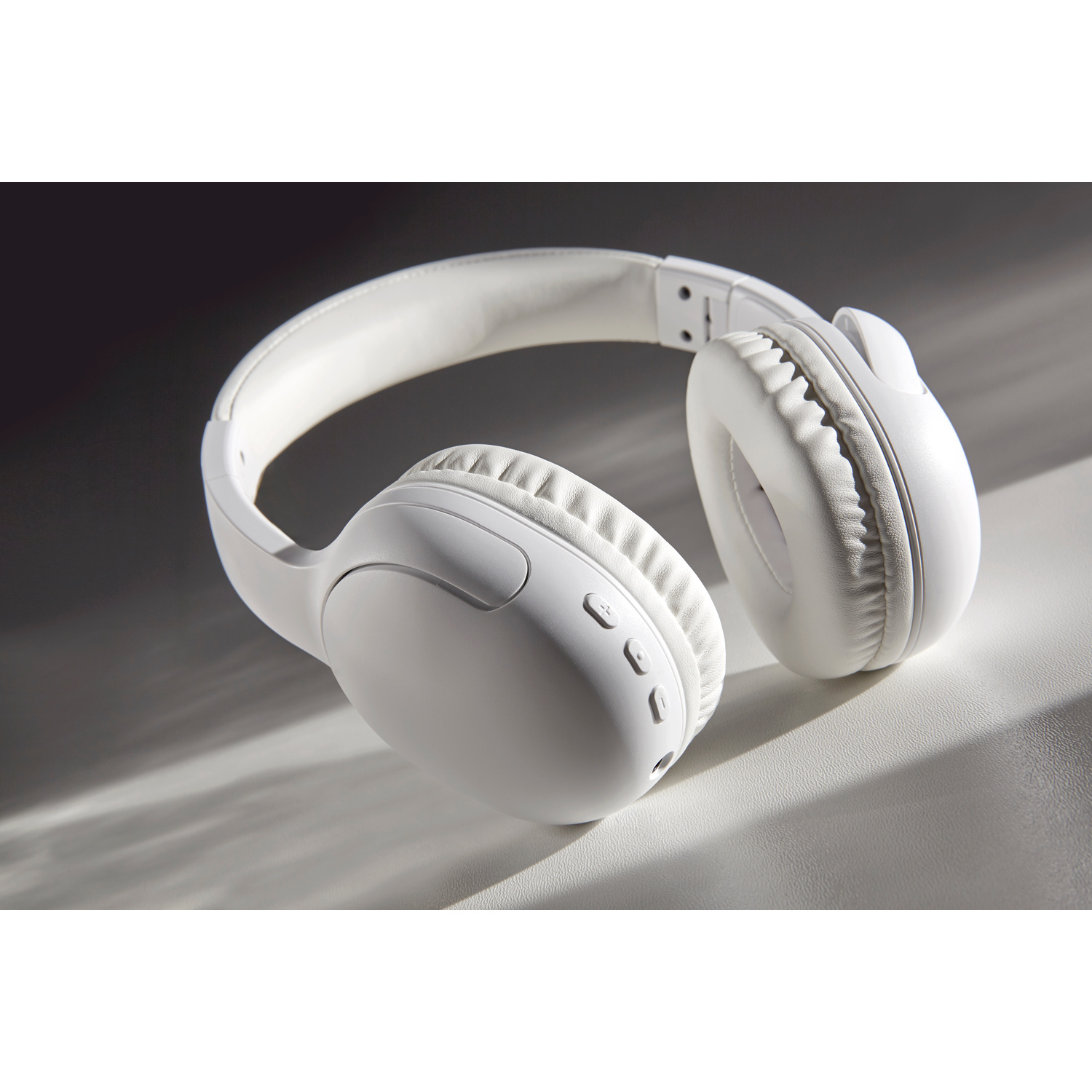 Un paio di cuffie bianche over-ear poggia su una superficie bianca testurizzata con ombre drammatiche.