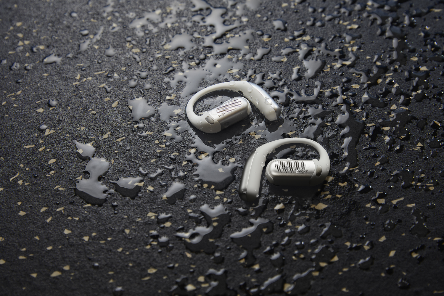 Dos auriculares inalámbricos blancos con ganchos para la oreja sobre una superficie oscura y húmeda.