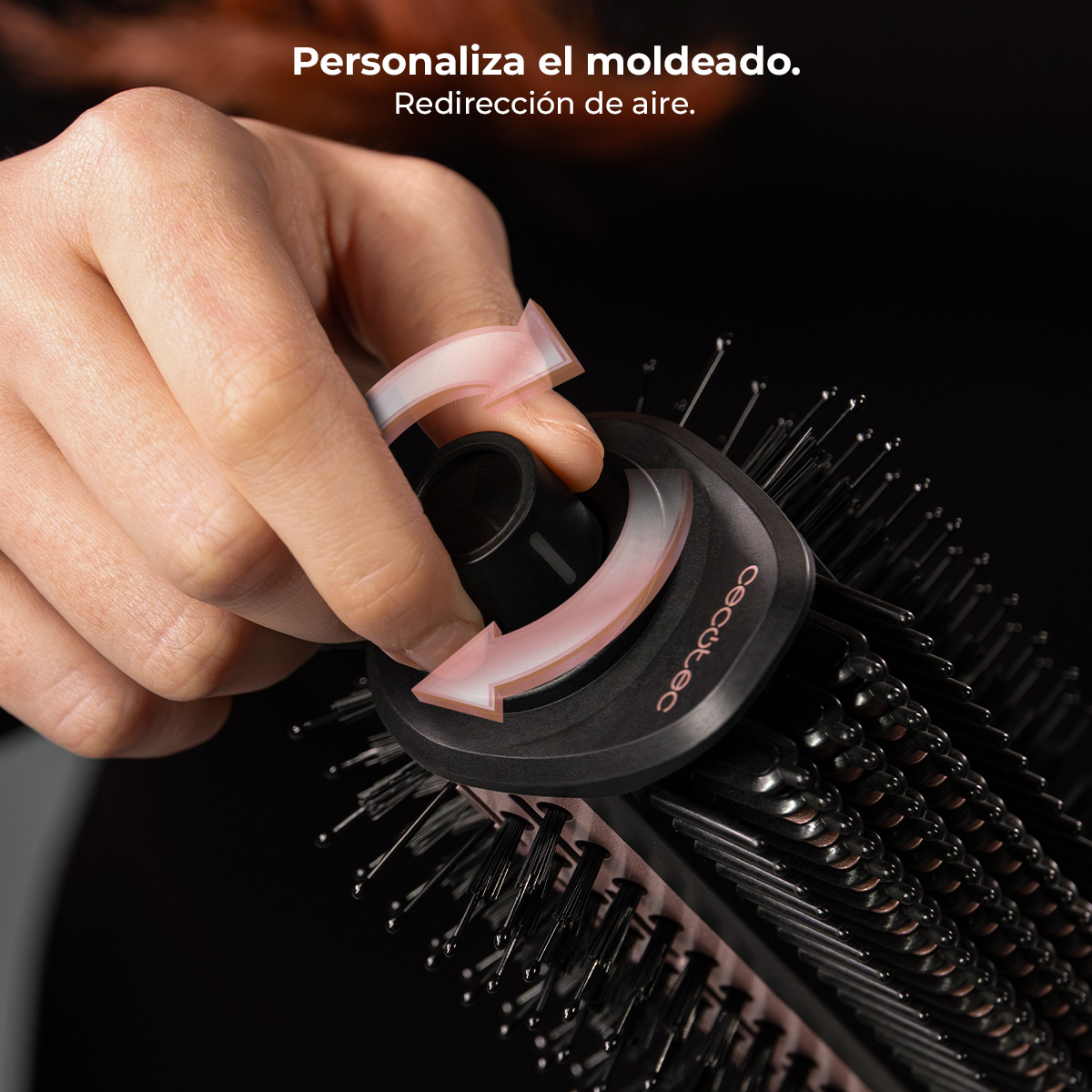 Una mano gira un mando negro en un accesorio de rizador con cerdas. Flechas rosas indican rotación.