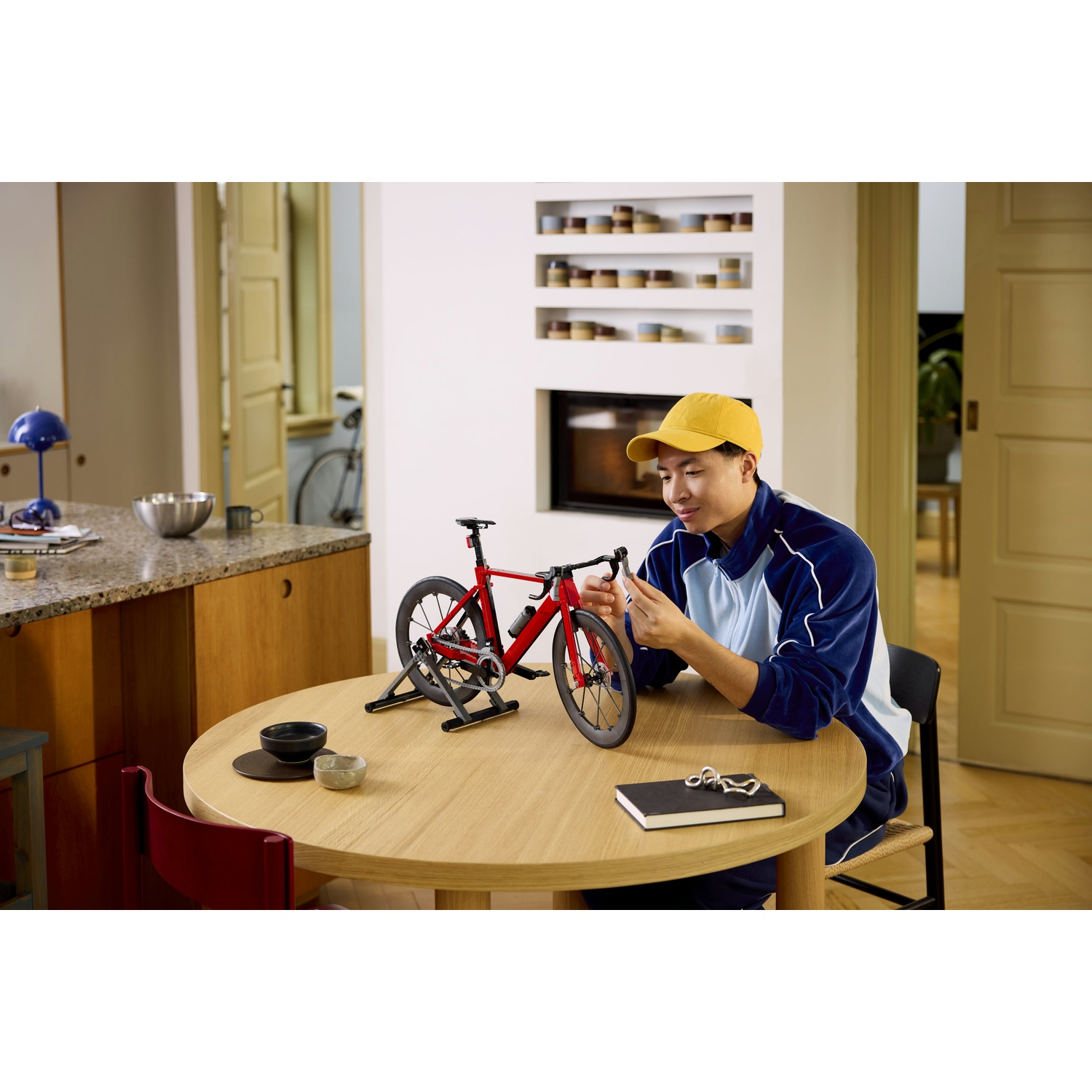 Uomo con cappellino giallo monta modello bici rossa su tavolo legno.