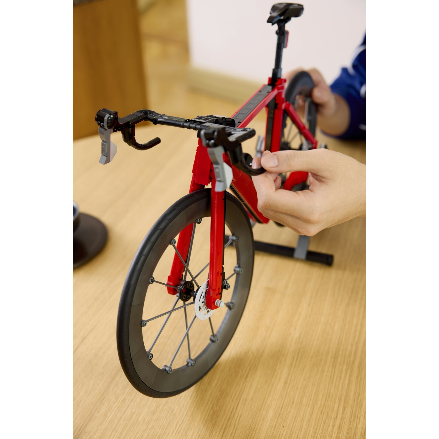 Una bicicletta da corsa Lego rossa con ruote nere su un tavolo di legno. Mani che la assemblano.