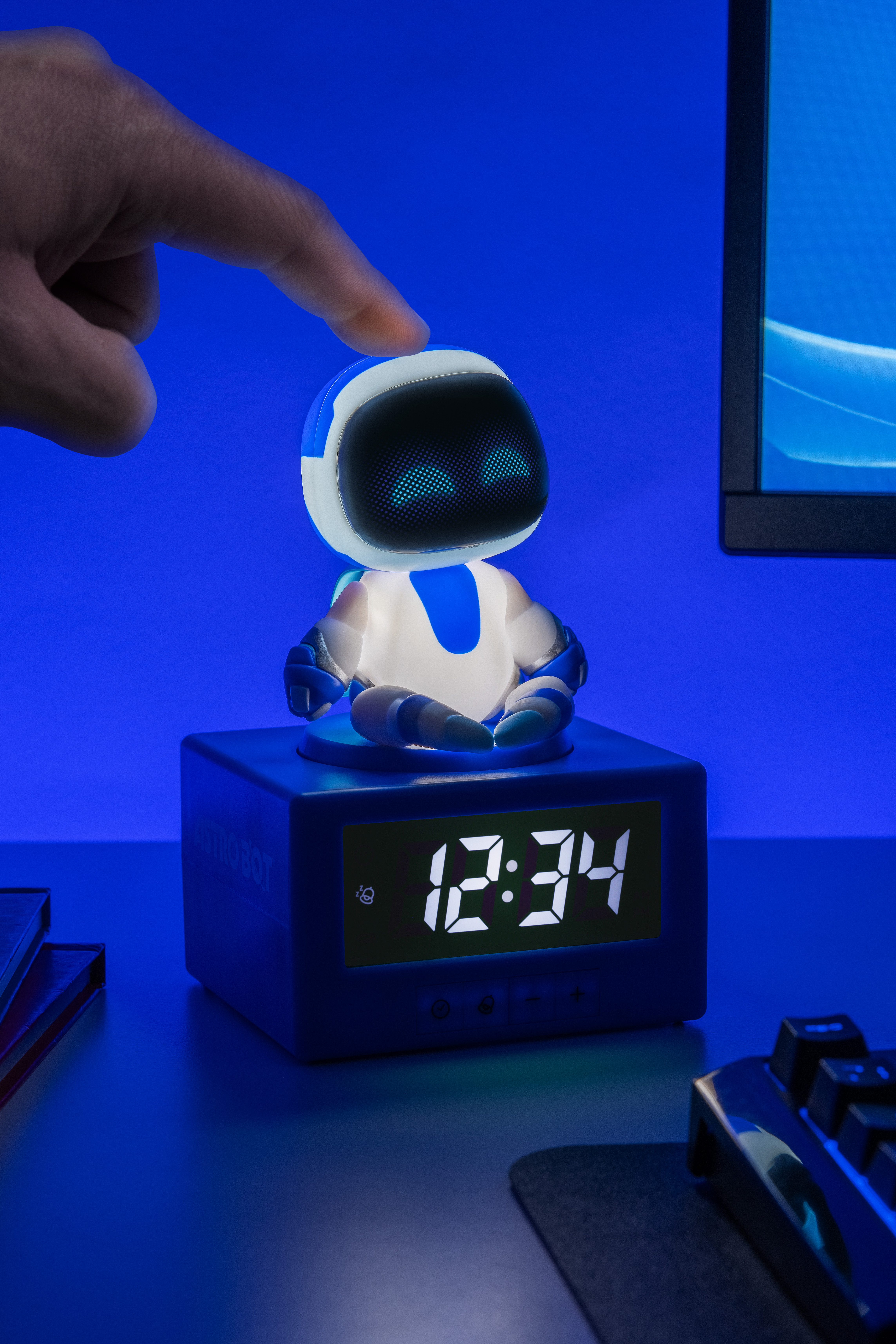 Ein Finger berührt einen beleuchteten Roboter auf einem blauen Wecker mit digitaler Anzeige.