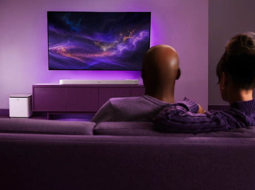 Két ember néz tévét lila hangulatfényben. Hangsáv és mélynyomó található.