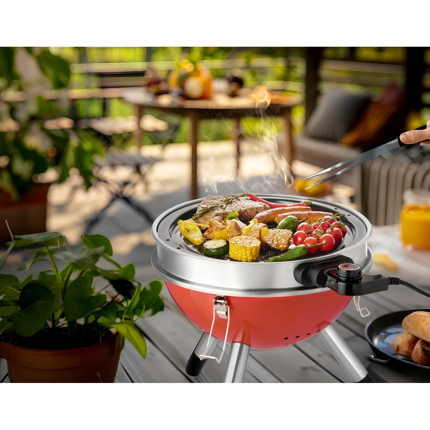 Rode elektrische grill met vlees, worstjes, groenten en kerstomaatjes op een houten terras.