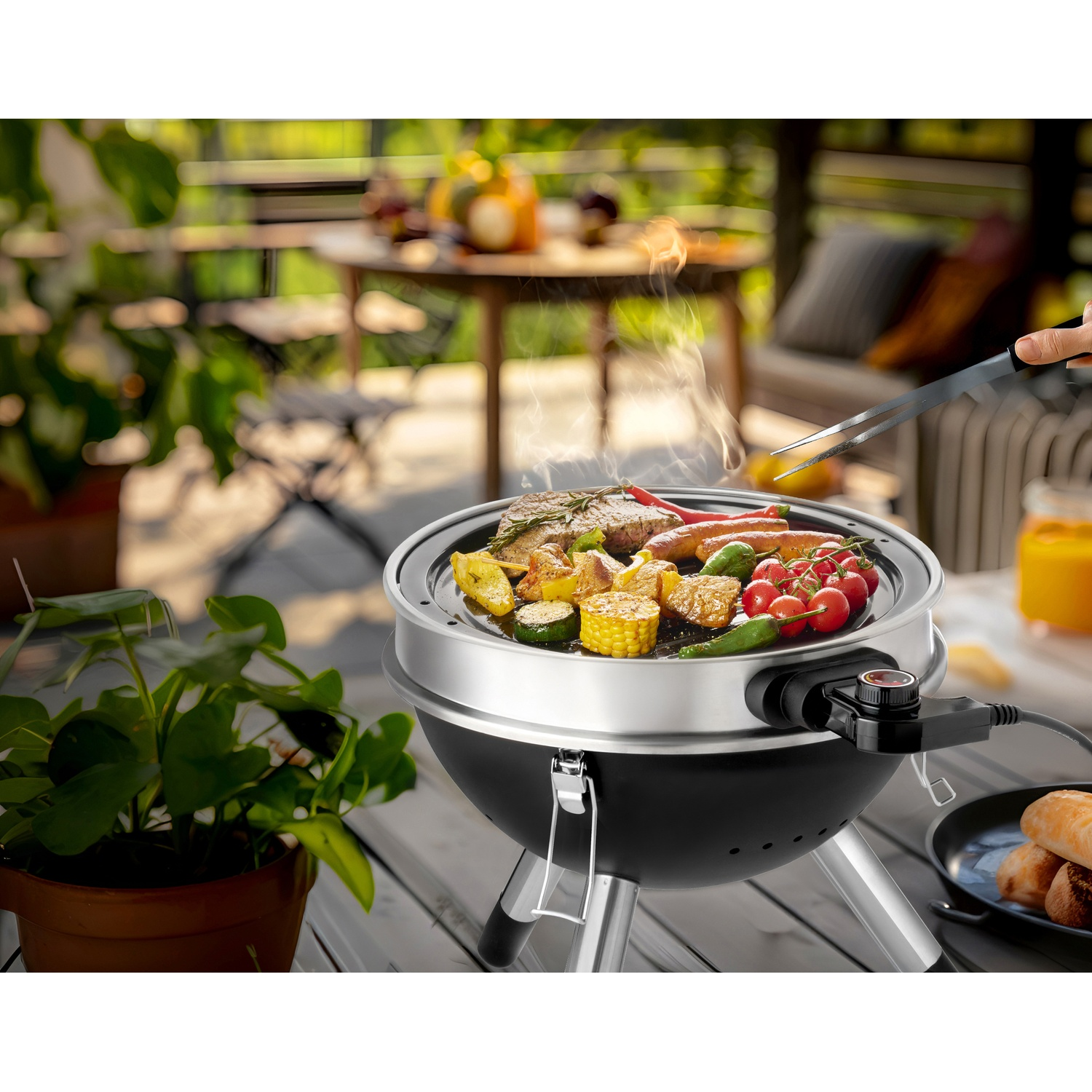 Electric grill with steak, sausages, vegetables, and tomatoes on a patio.