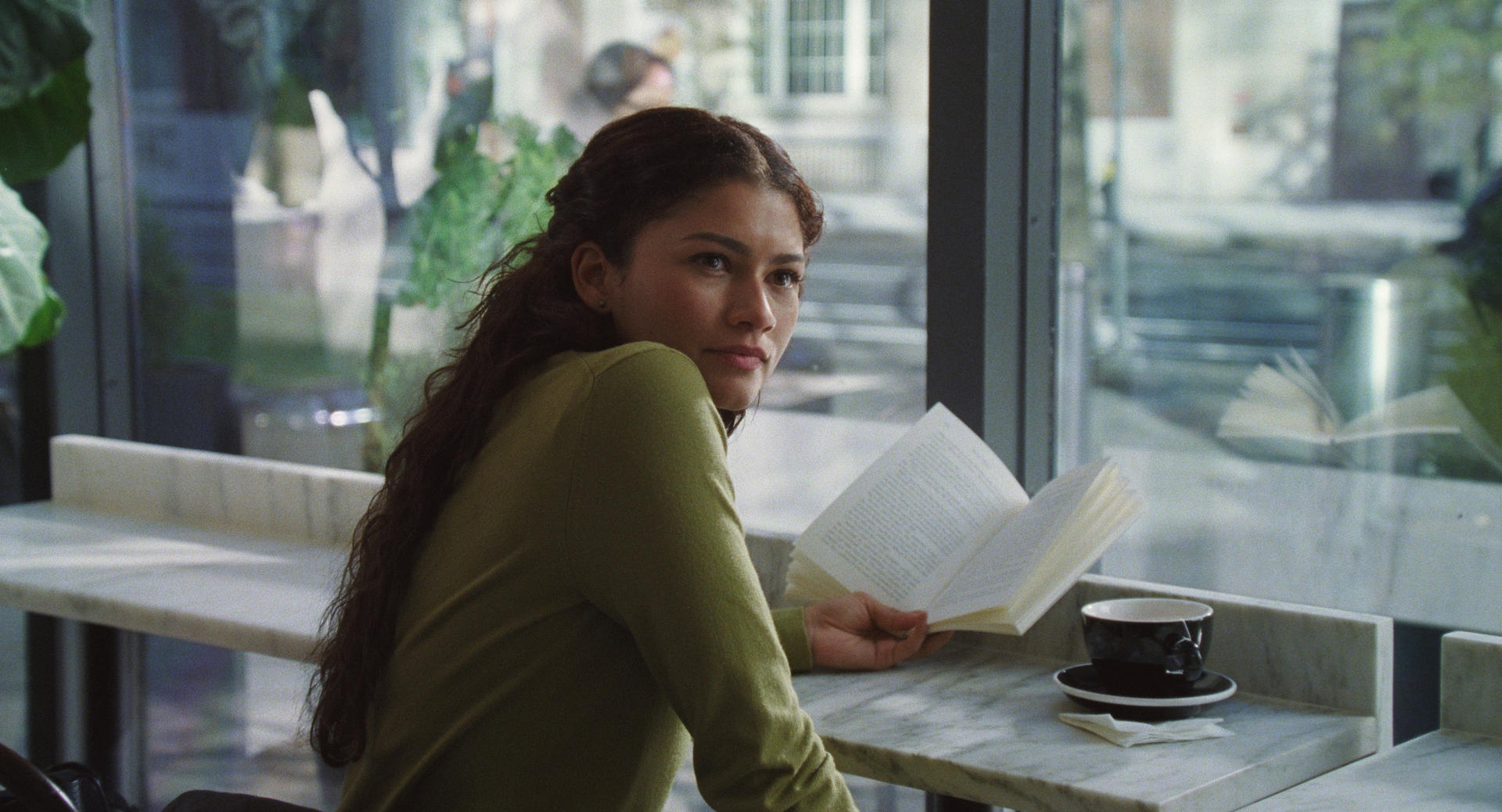 Eine Frau mit dunklem Haar liest ein Buch in einem Café mit Blick nach draußen.